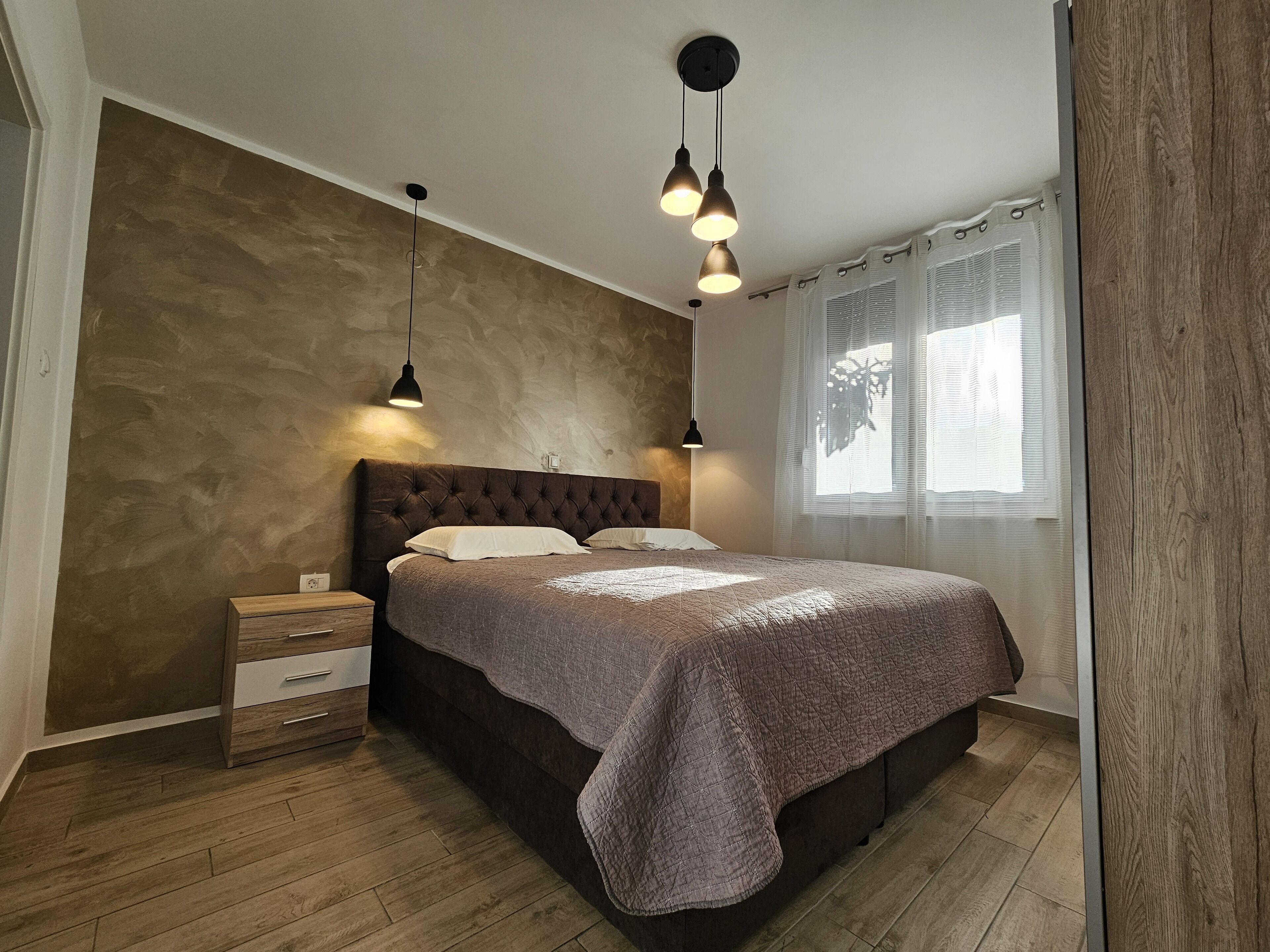 Un dormitorio con cabecera capitonada, mesita de noche de madera, cama con cubrecama acolchado e iluminación colgante.