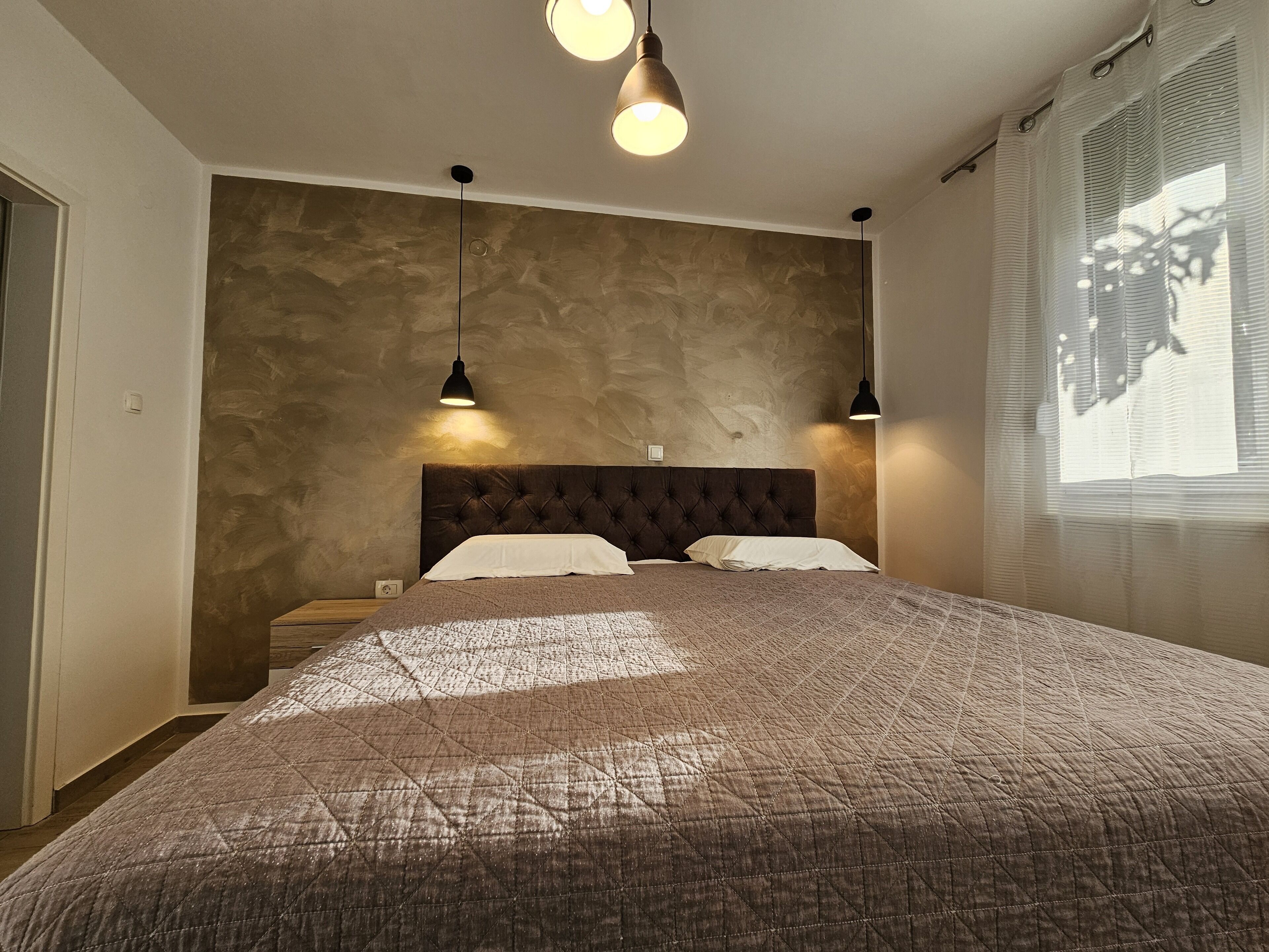 Un dormitorio con una cama grande, dos lámparas colgantes, una pared con textura y una ventana con persianas.