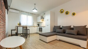 A modern living room with a sectional sofa, a small dining table, and a kitchen area with white cabinets and a hanging light fixture. - Deluxe Apartment near Bridge (Zadar)