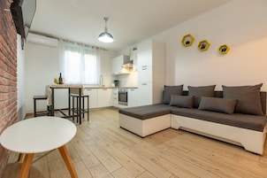Ein modernes Wohnzimmer mit einer Eckcouch, einem kleinen Esstisch und einer Küchenzeile mit weißen Schränken und einer Pendelleuchte.