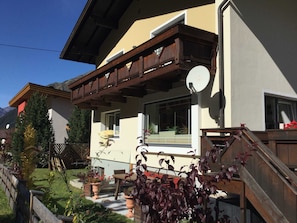 A two-story house with a balcony, a satellite dish, and a garden with various plants and a bench.