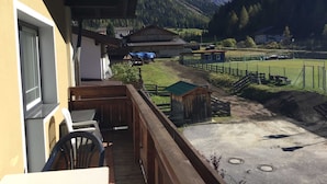 Ein Balkon mit Blick auf eine gebirgige Landschaft, ein eingezäuntes Sportfeld und eine kleine Holzkonstruktion.
