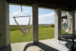Een hangmat bevestigd aan een betonnen pilaar, een ronde tafel met een tafelkleed met dessin, en uitzicht op een grasveld door een raam.