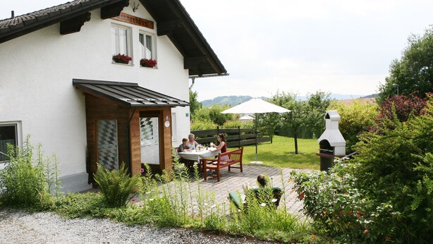 A white building with a wooden door and window, a bench, and a barbecue grill in the garden. - Cozy 3-star vacation home, quiet and close to nature for 4 people / 2 bedrooms (Zwiesel)