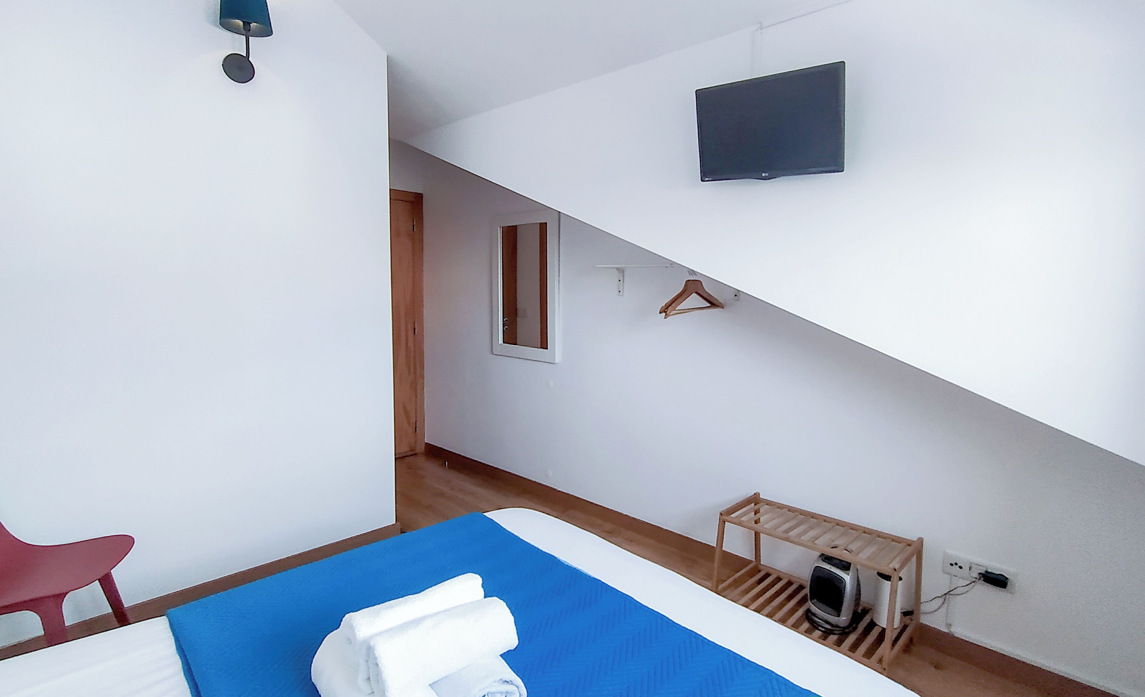 A hotel room with a bed, a TV mounted on the wall, a small wooden shelf, and a red chair.