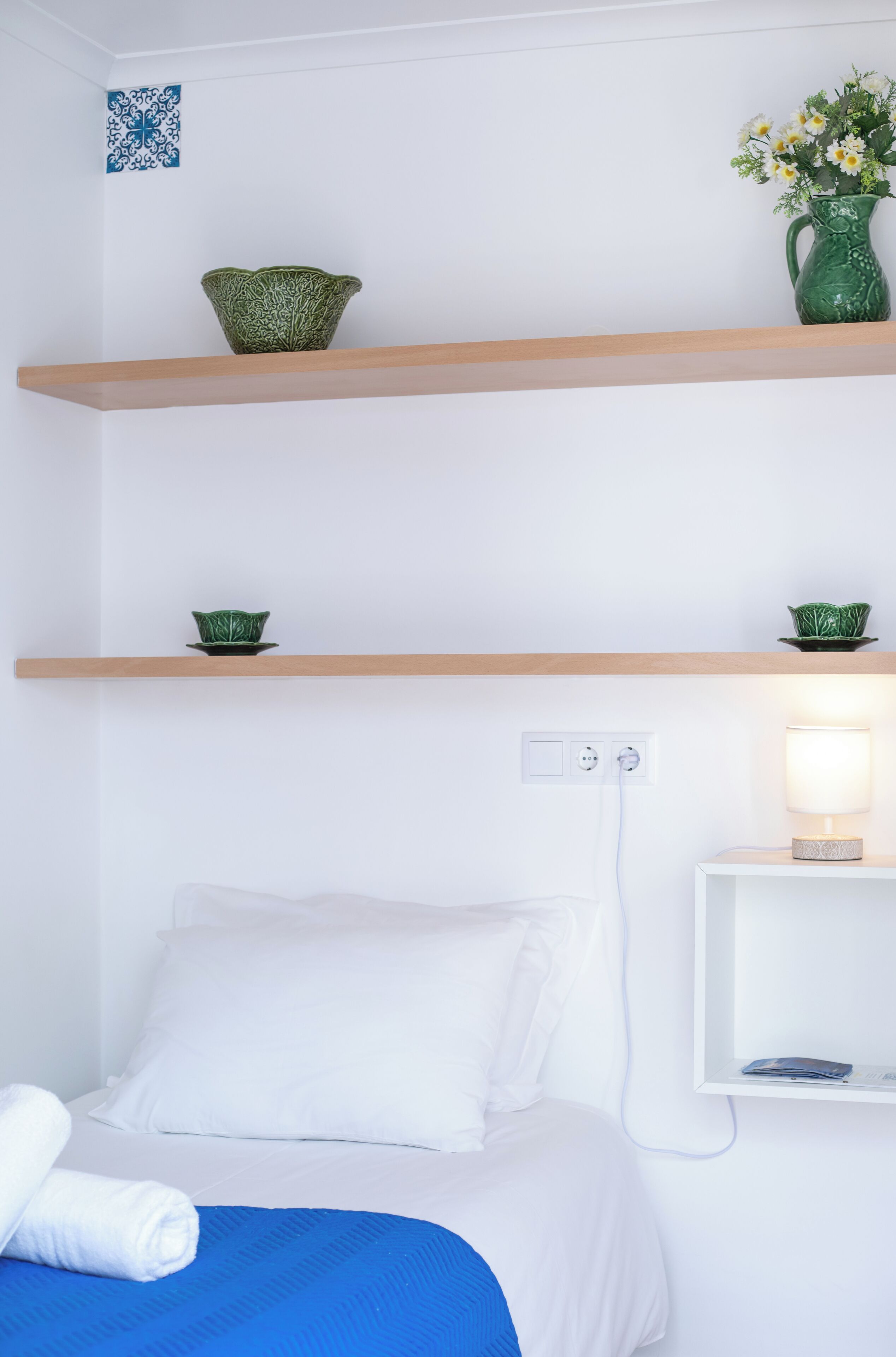Quarto de hotel com cama, manta azul, almofadas brancas, vaso verde com flores e prateleira de madeira com objetos decorativos.