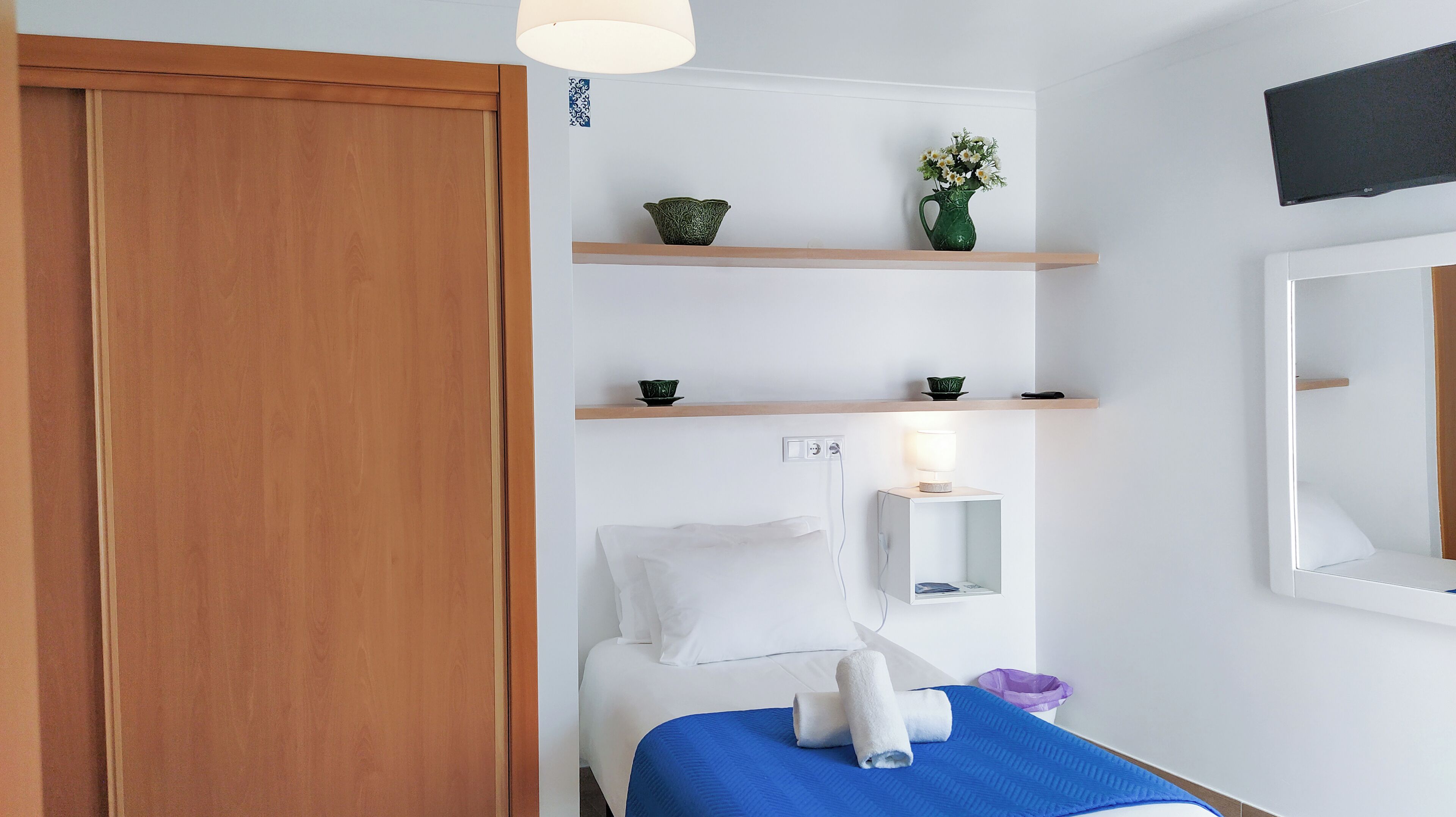 Habitación de hotel con un armario de madera, una cama con ropa blanca y un plaid azul, una mesita de noche con una lámpara, y un estante con objetos decorativos.