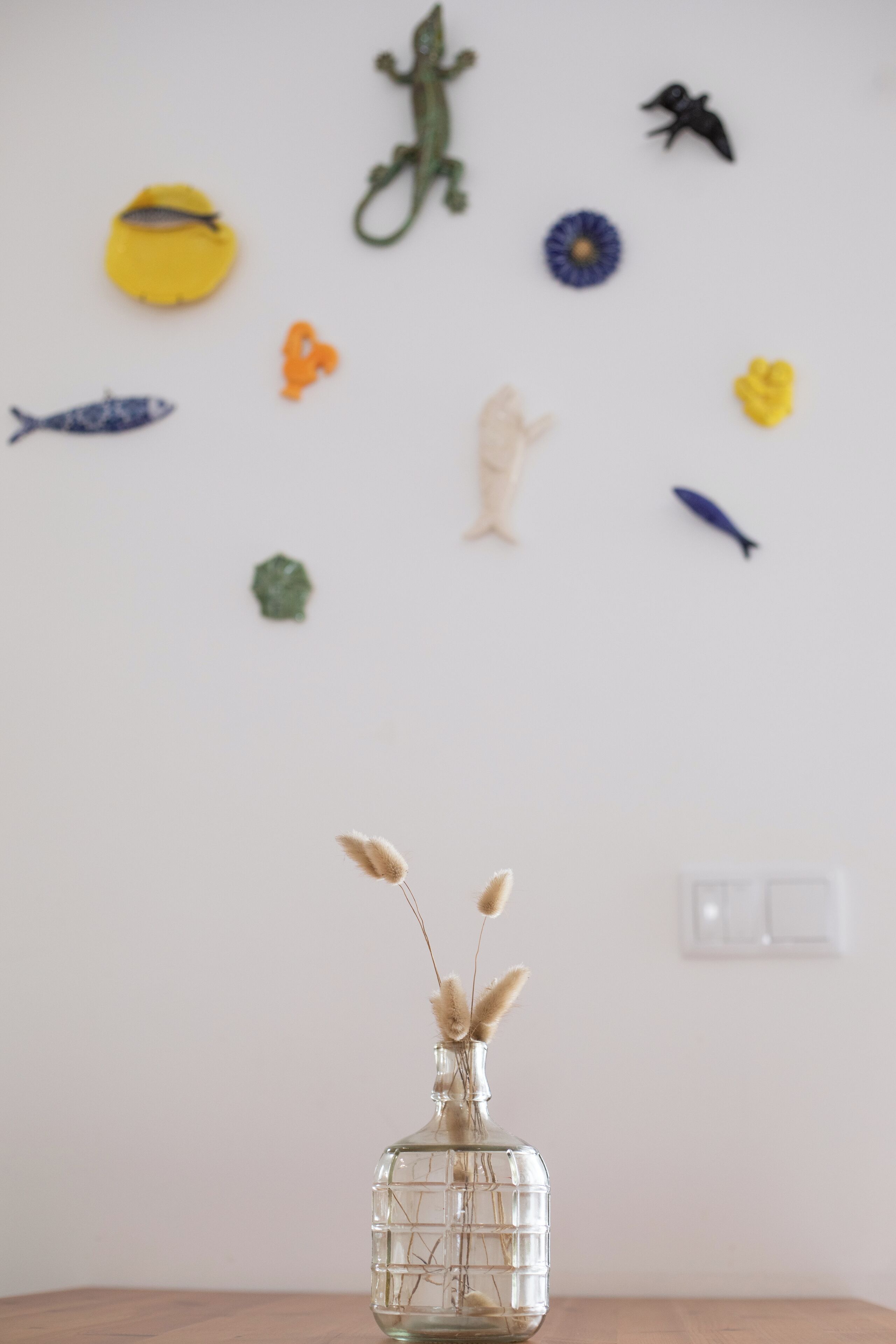 Un vase en verre transparent contenant des plantes séchées, posé sur une table en bois devant un mur blanc, agrémenté de divers petits objets décoratifs.