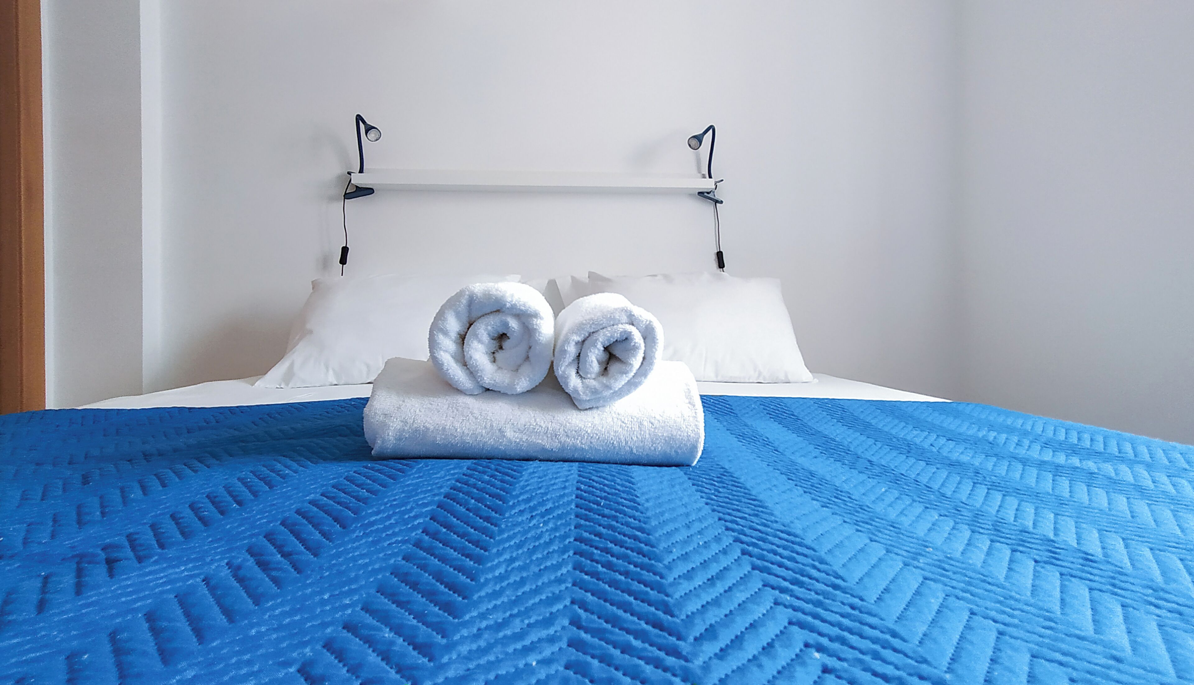 Habitación de hotel con cama, colcha azul con textura, dos toallas enrolladas, almohadas blancas y pared blanca.