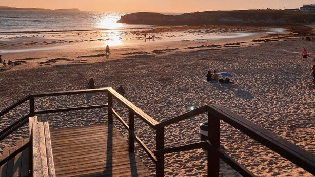Una playa con escaleras de madera que bajan a la arena, cielo despejado y el sol poniéndose sobre el océano.