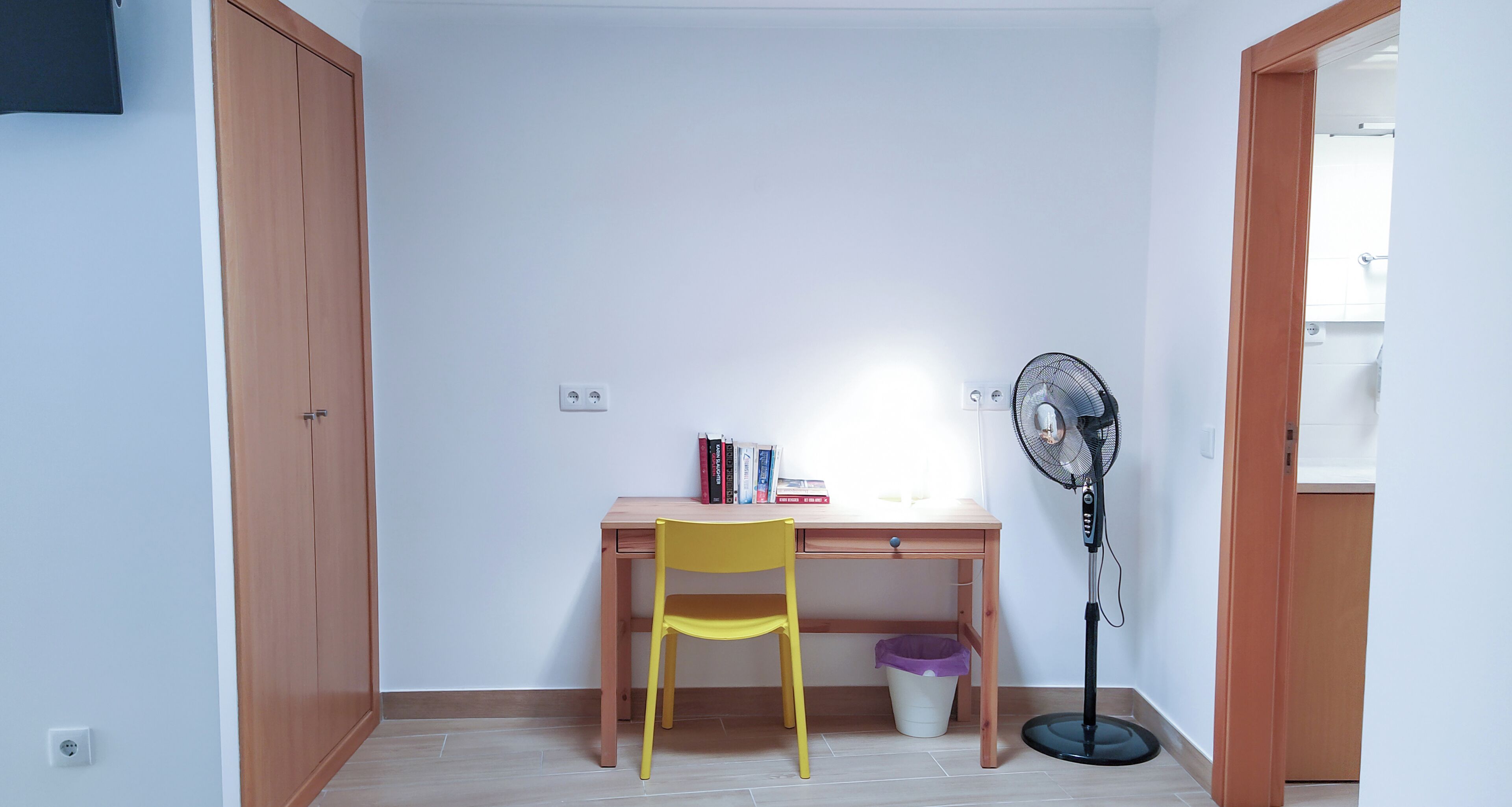 Una habitación con un escritorio de madera, una silla amarilla, un ventilador de pie y un cesto de basura.