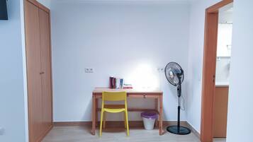 Una habitación con un escritorio de madera, una silla amarilla, un ventilador de pie y un cesto de basura.