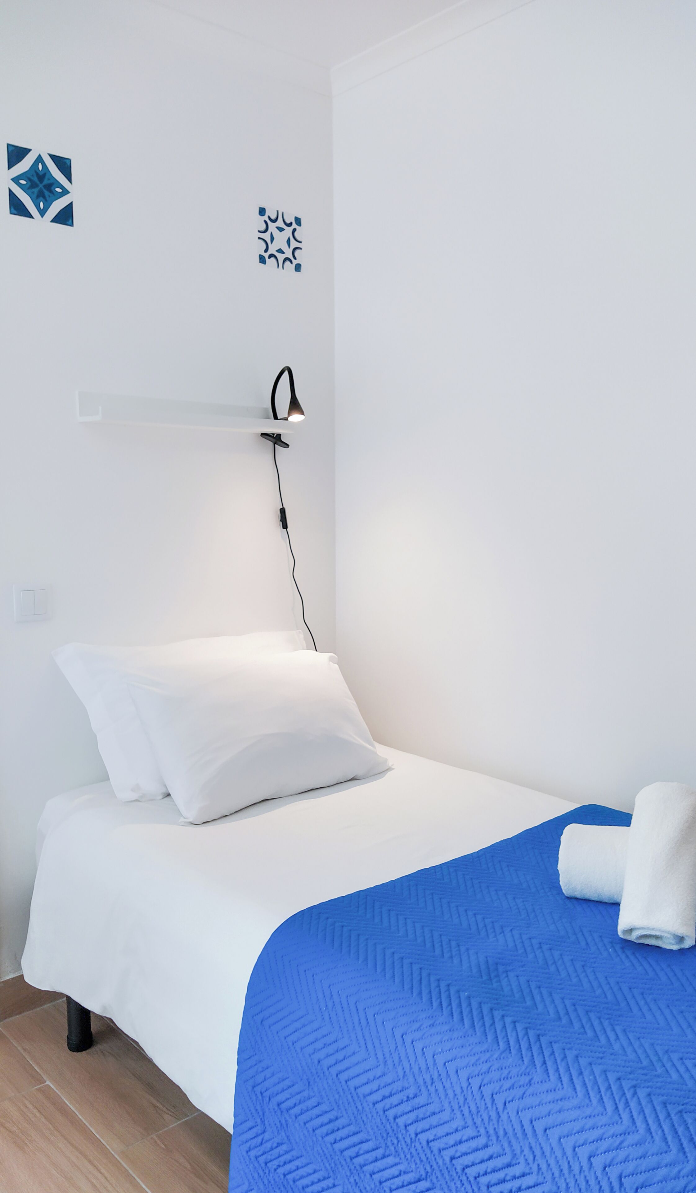 A neatly made bed with white bedding and a blue blanket, a bedside table with a lamp, and a wall-mounted power outlet.