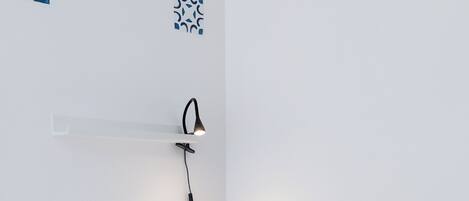 A neatly made bed with white bedding and a blue blanket, a bedside table with a lamp, and a wall-mounted power outlet.