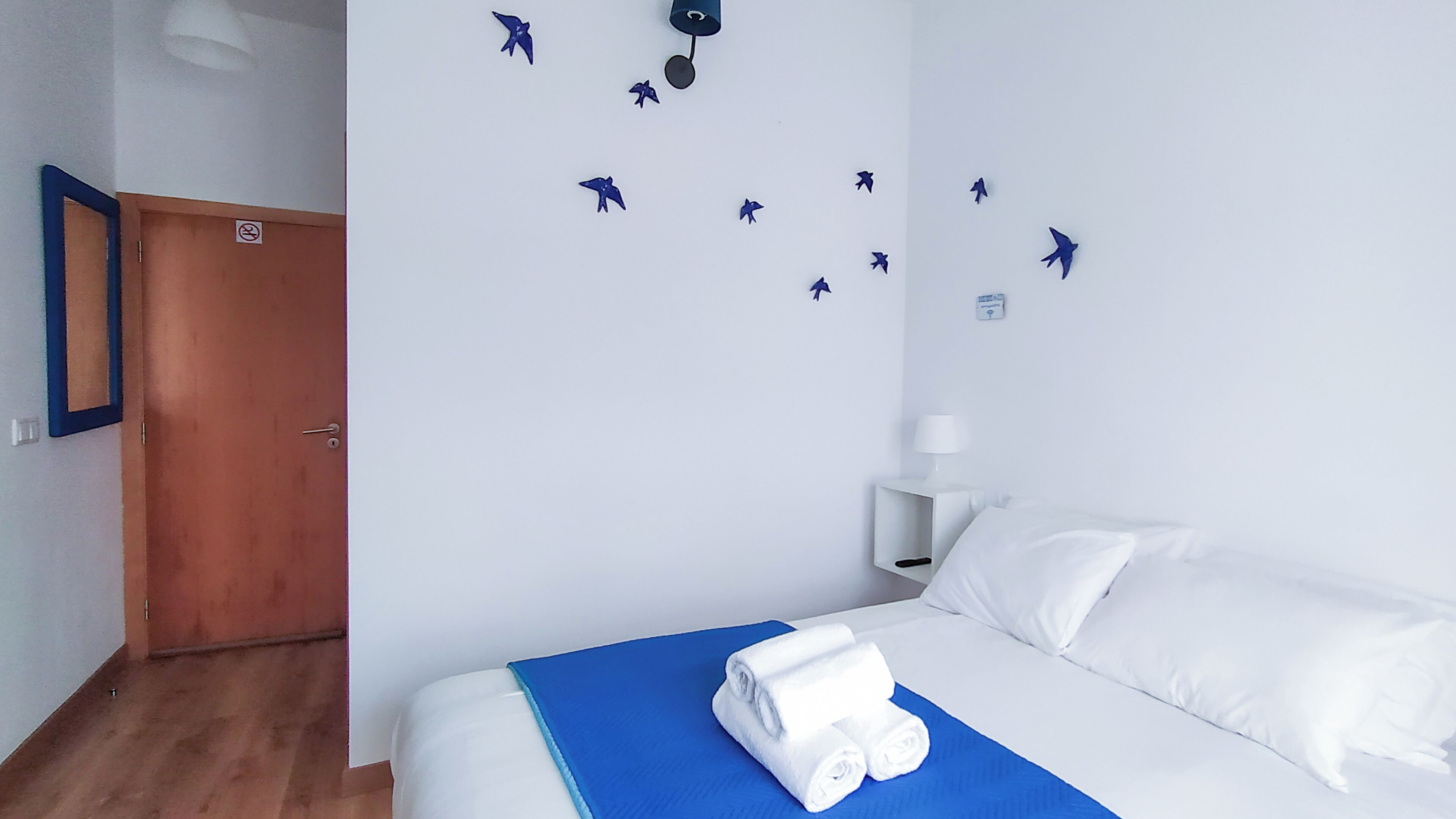 A hotel room with a bed, a blue blanket, white pillows, a wooden door, and a wall with bird decals.