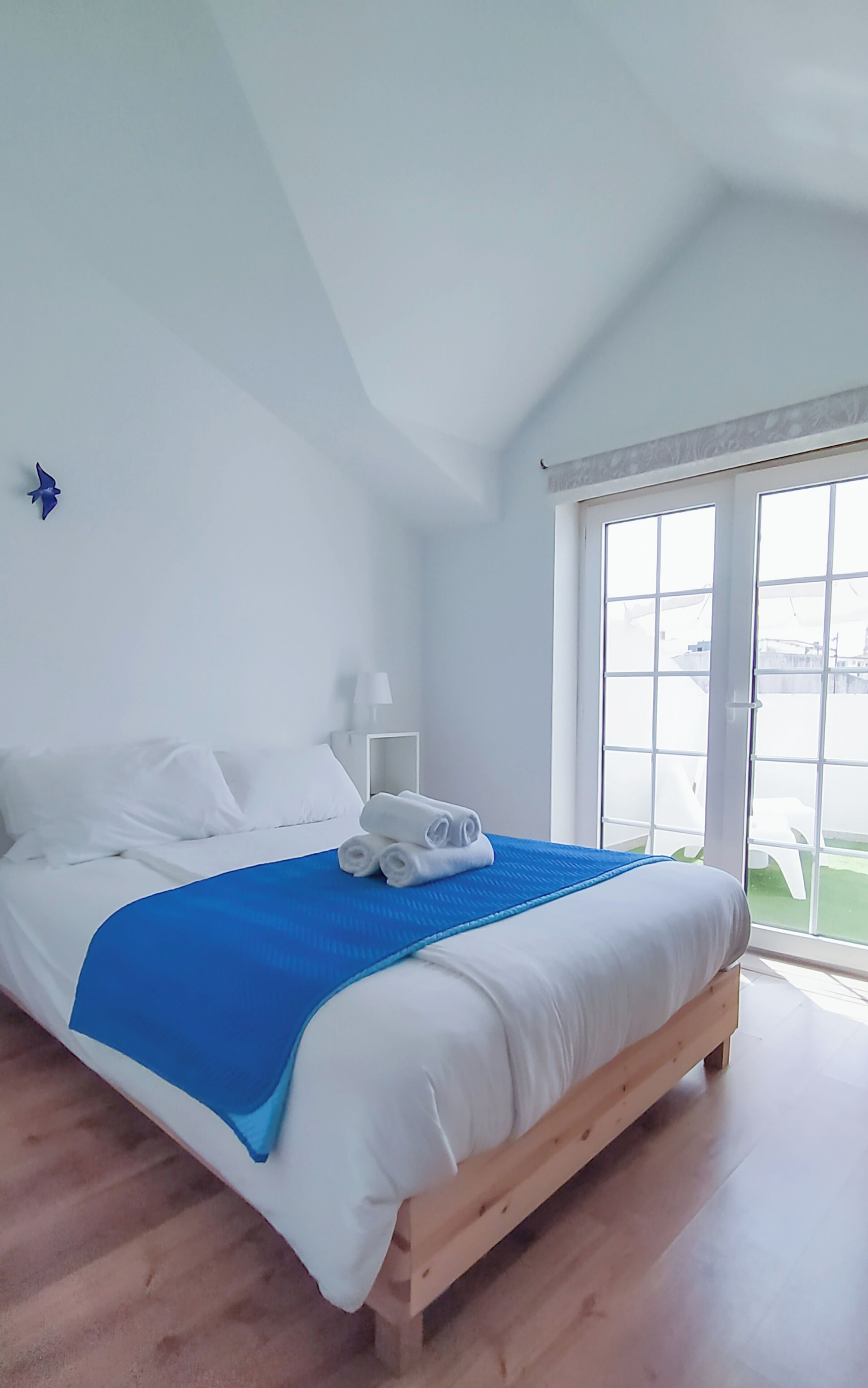 A bedroom with a bed, white linens, a blue accent, a nightstand, a lamp, and a window with a view.