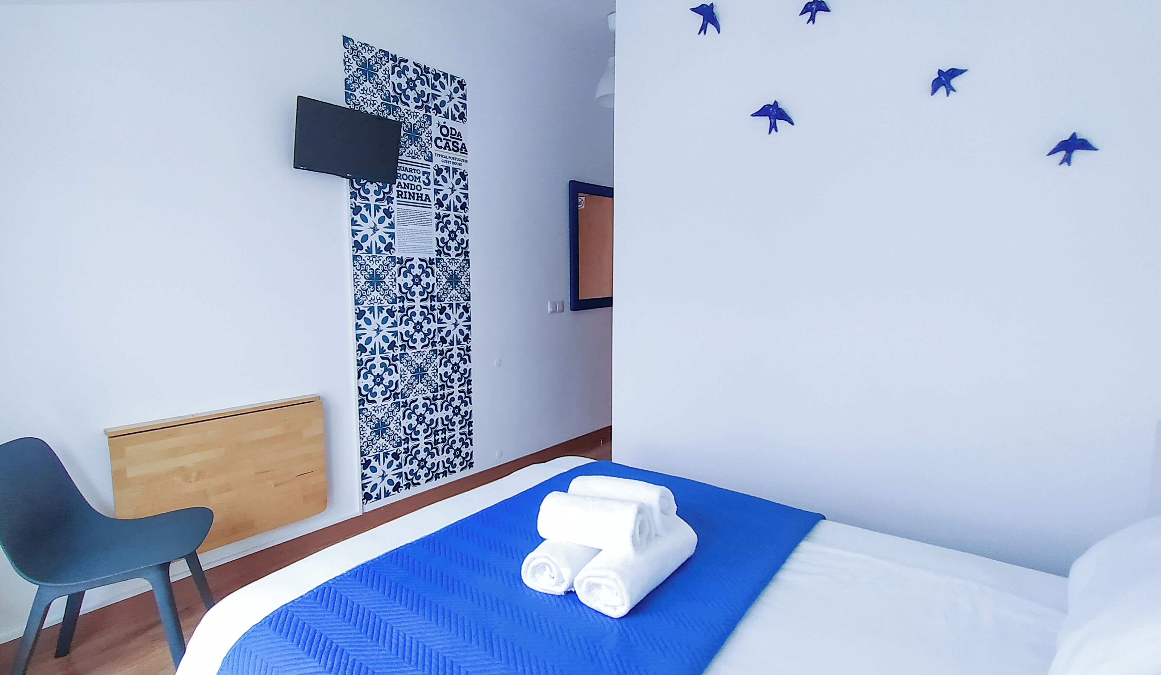 Habitación de hotel con una cama, un edredón azul, una mesita de noche de madera, una silla negra, un papel tapiz estampado, un televisor de pantalla plana y una obra de arte en la pared con aves.