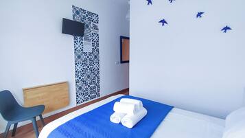 Habitación de hotel con una cama, un edredón azul, una mesita de noche de madera, una silla negra, un papel tapiz estampado, un televisor de pantalla plana y una obra de arte en la pared con aves.