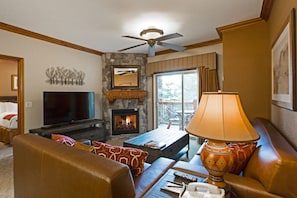 A living room with a stone fireplace, a television, a ceiling fan, and a large window.
