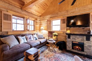 A log cabin interior with a leather sofa, a flat-screen TV, a stone fireplace, and a large window.
