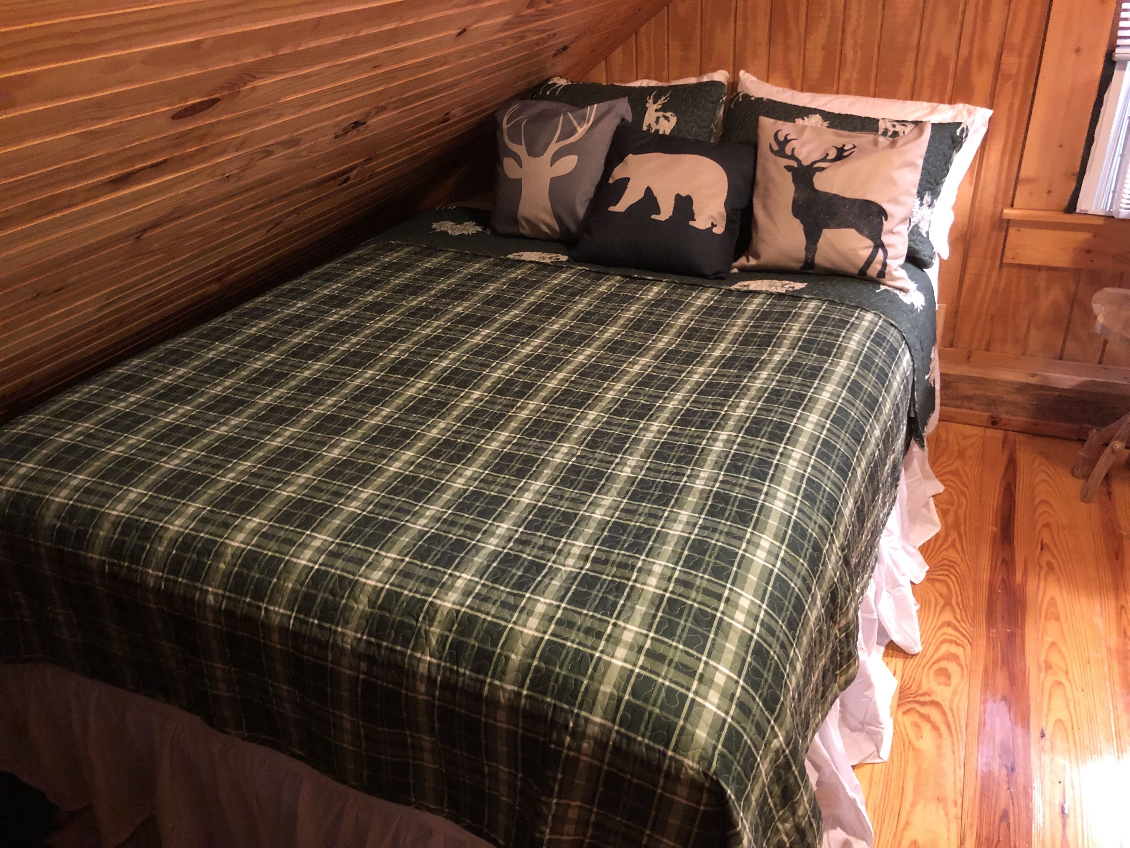 Une chambre dans une cabane en bois, avec une couvre-lit à carreaux, un coussin orné d’un motif de cerf et un plafond en bois.