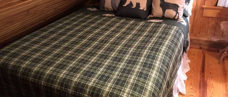 Une chambre dans une cabane en bois, avec une couvre-lit à carreaux, un coussin orné d’un motif de cerf et un plafond en bois.