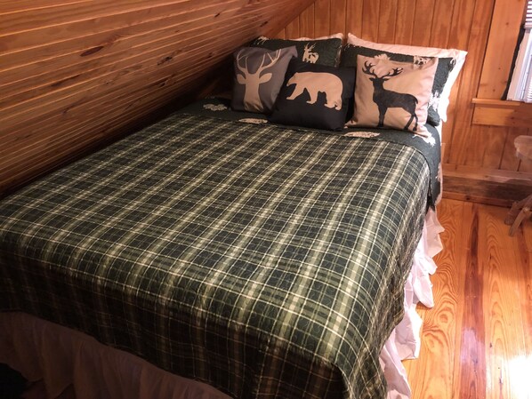 Une chambre dans une cabane en bois, avec une couvre-lit à carreaux, un coussin orné d’un motif de cerf et un plafond en bois.