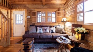 A cozy log cabin interior with a wooden ceiling, a sofa with decorative pillows, a small table with a lamp, and a window with shutters. - Shaded Log Cabin Swim Spa 2 miles Lake Entrance & winery (Birdseye)