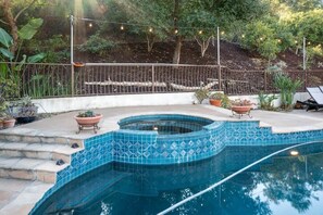 A swimming pool with blue mosaic tiles, surrounded by a stone deck and potted plants.