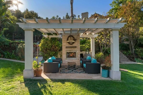 A covered outdoor seating area with a stone fireplace, surrounded by potted plants and a pergola.
