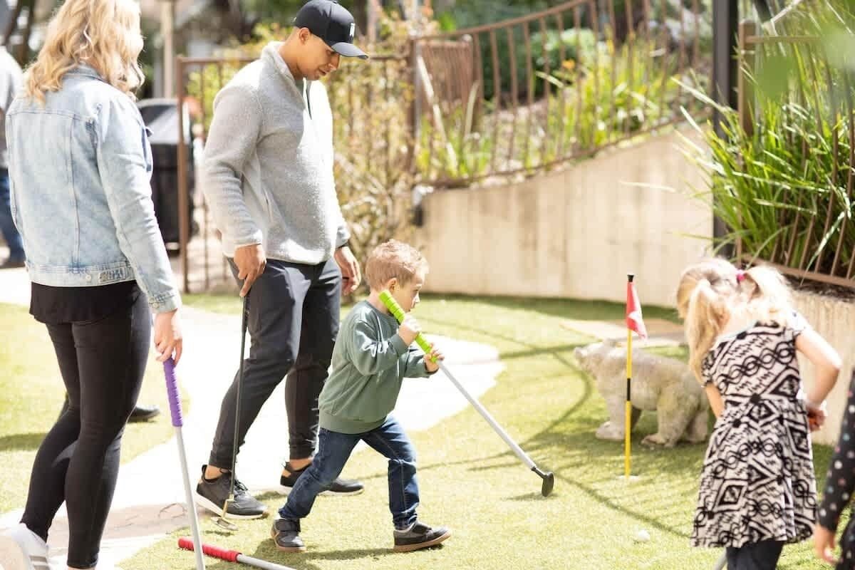A group of people playing croquet in a backyard with a lawn, a stone statue, and a flagpole.