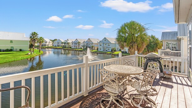 Un balcon donnant sur le front de mer, avec une table et des chaises, une balustrade blanche et une vue sur les maisons de l’autre côté du canal.