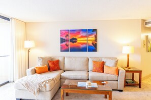 En stue med en beige sofa, to orange puder, et glas sofabord og et maleri af en solnedgang over vandet.