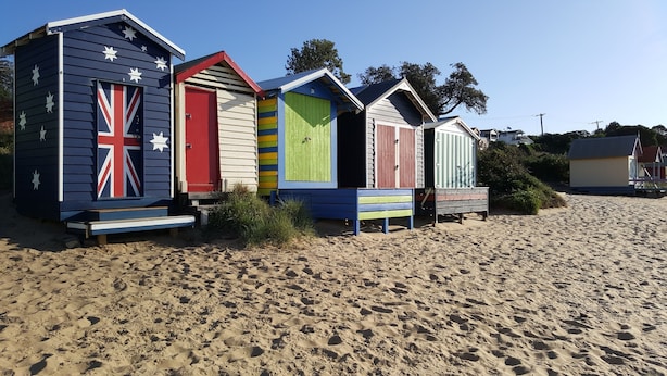 A row of beach huts with varying colors and designs, including a Union Jack motif, on a sandy beach.