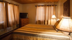 Habitación de hotel con techo de madera, cama con ropa de cama a rayas, televisor sobre una cómoda de madera y lámpara en una mesita de noche.
