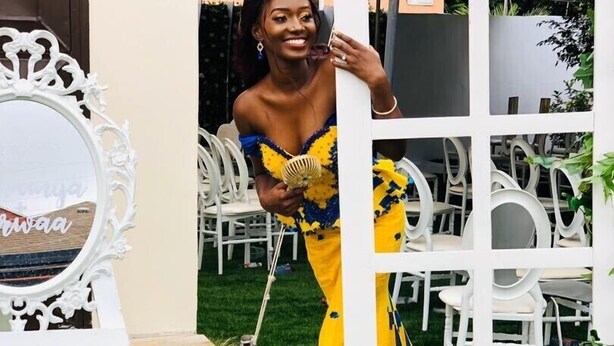 Une femme, vêtue d’une robe jaune et bleue, se tient devant un portail blanc.