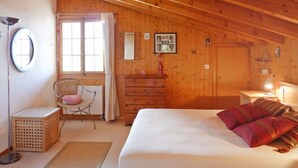 Een gezellig slaapkamer met een houten plafond, een bed met witte beddengoed en rode kussens, een houten dressoir, een stoel met een roze kussen, een klein tafeltje met een lamp, en een raam met gordijnen.