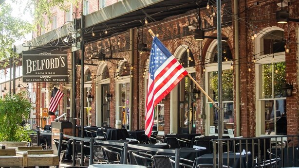 An outdoor seating area with tables and chairs, a brick building facade, and an American flag. - Amazing Loft with Balcony Overlooking River Street by Lucky Savannah (Savannah)