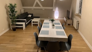 A modern living room with a wooden floor, a black sofa, a white coffee table, and a dining table with chairs. There is a potted plant and a radiator on the wall.