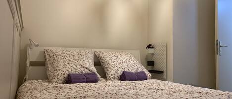 Un dormitorio con edredón estampado con motivos florales, dos almohadas, una mesita de noche con una planta y un aplique de pared.