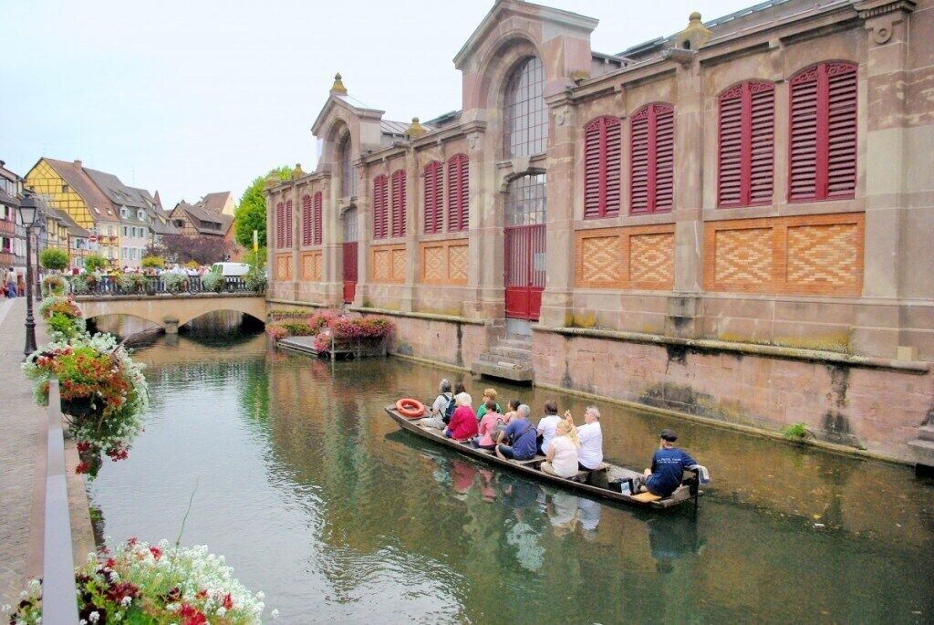 A canal with a boat carrying passengers, surrounded by historic buildings with red shutters and a stone bridge in the background.