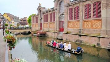 A canal with a boat carrying passengers, surrounded by historic buildings with red shutters and a stone bridge in the background.