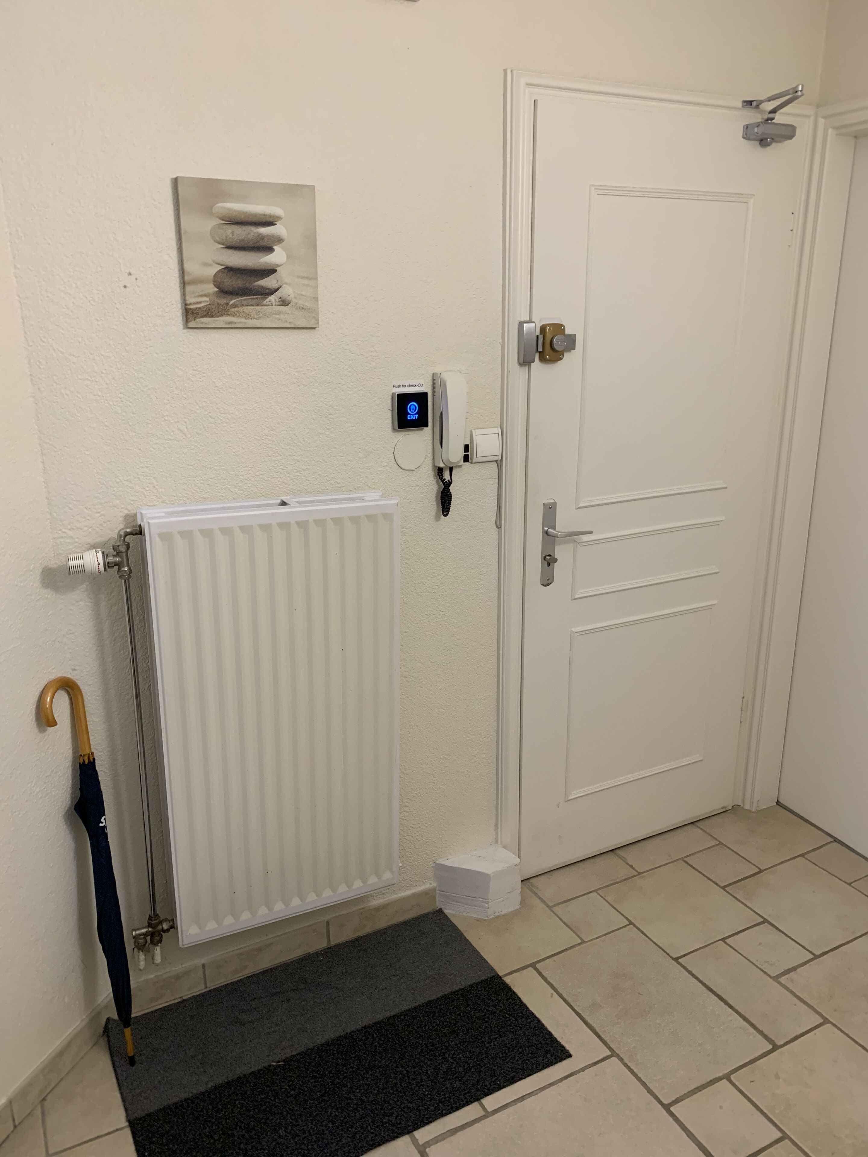 Una habitación con un radiador blanco, una puerta con mirilla, un portero automático con botón pulsador y un paraguas azul apoyado en el radiador.