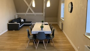A modern loft apartment with exposed wooden beams, a dining area with a wooden table and chairs, a black sofa, and a large window providing natural light.