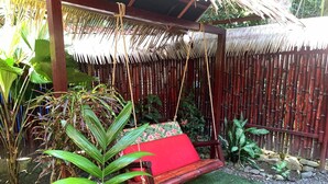 Una hamaca de madera con cojín rojo, rodeada de exuberante follaje y una valla de bambú.