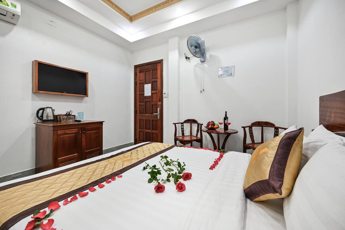 A hotel room with a bed, a wooden cabinet, a television, a fan, and a small table with a bottle and glasses.