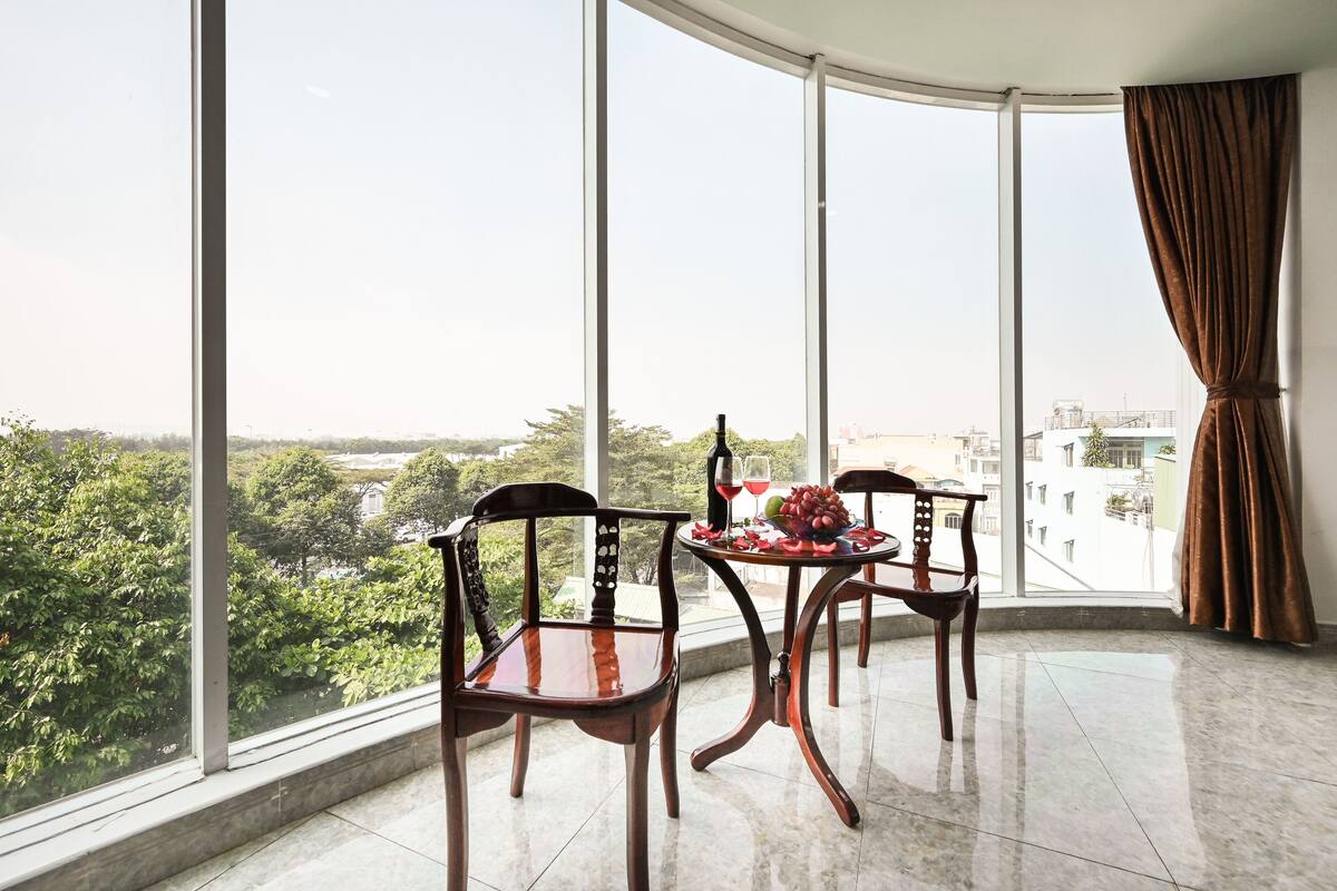 A balcony with two wooden chairs, a table with fruit and a glass of red wine, and a view of greenery and buildings.