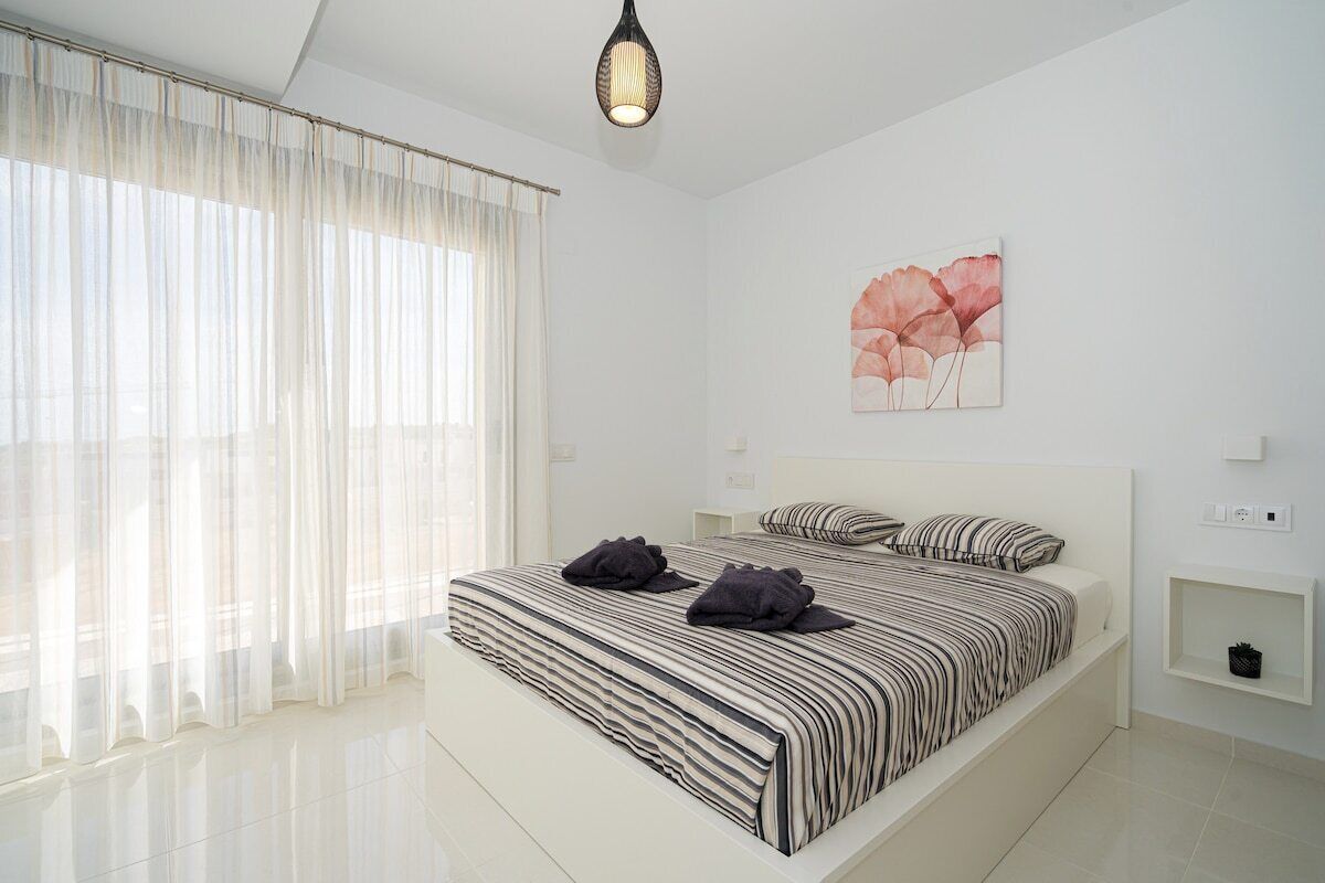 Une chambre à coucher moderne avec un grand lit, des rideaux légers, un luminaire fixé au mur et une œuvre d’art florale.