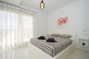 Un dormitorio moderno con una cama grande, cortinas translúcidas, una lámpara de pared y una obra de arte floral.