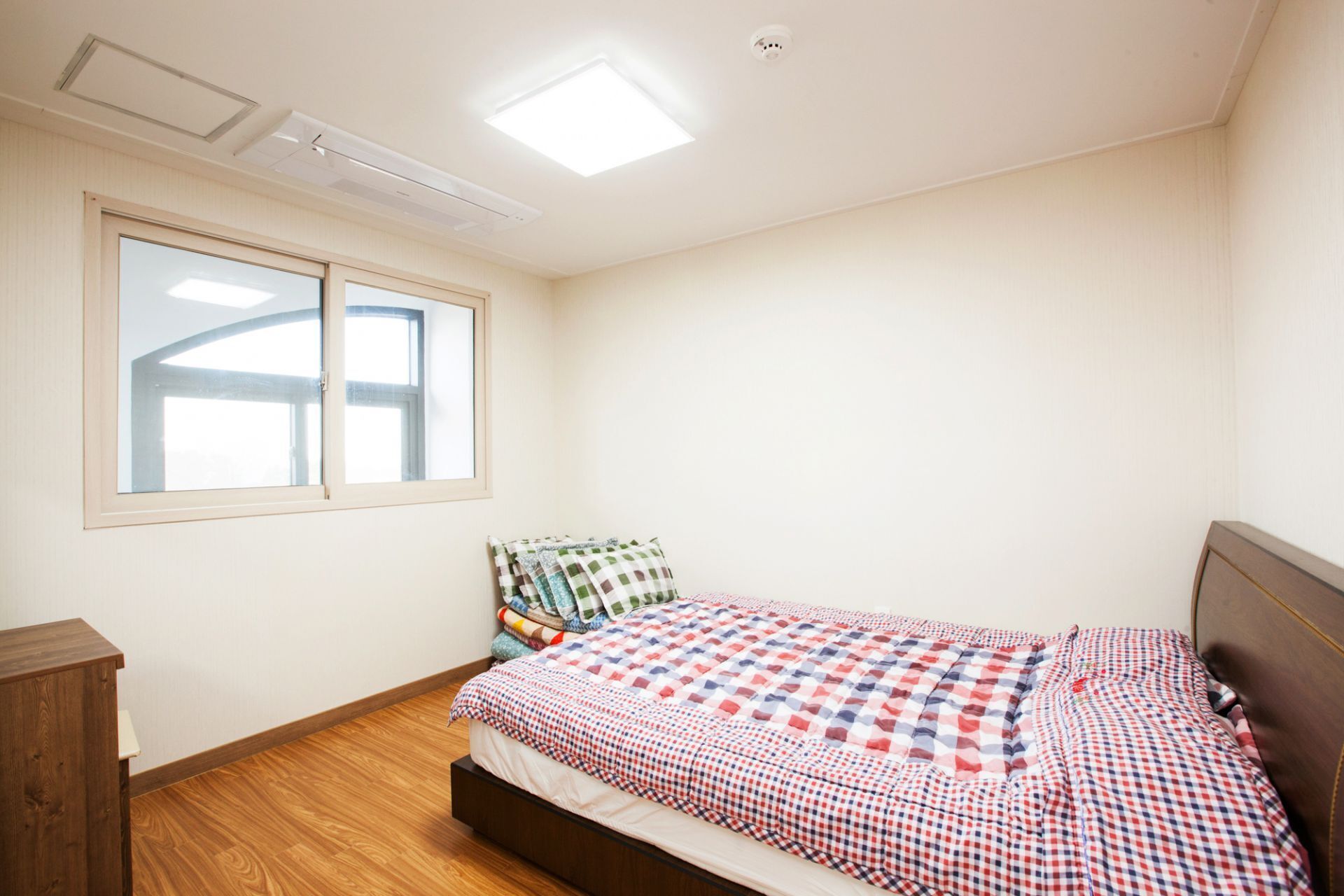 A bedroom with a bed, a wooden nightstand, a window, and a ceiling light.